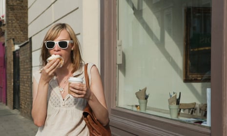 A young woman in the street eating what looks like a cupcake, and holding a cup of coffee.