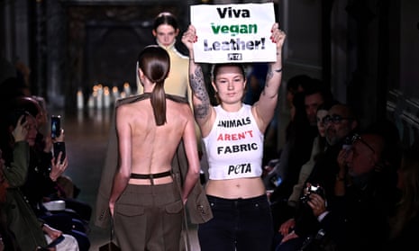 Protester holding a placard reading ‘Viva vegan leather!’ and wearing a vest top reading ‘Animals aren’t fabric’, with catwalk models in the background.