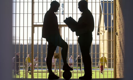 A man with a clipboard and a younger man with a football converse silhouetted against a barred gate.