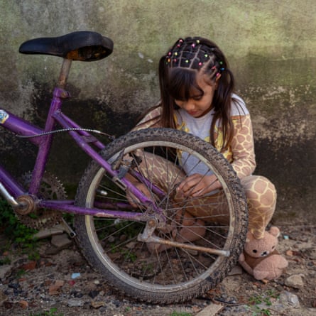 A small girl squats amid silt and small stones, with a wall behind her that is green with algae, as she puts the chain back on her bike