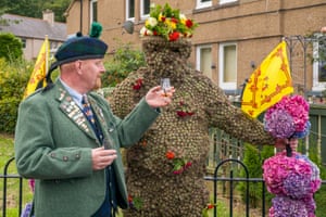 The man in traditional Scottish attire, swearing a green tweed jacket adorned with various pins and badges and a Glengarry hat with a tartan pattern.