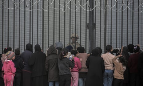 A crowd of what appears to be women and children is pictured from behind. In front of them is a vast metal fence with curled barbed wire running along the top. On the other side of the fence a lone Egyptian soldier faces the group.