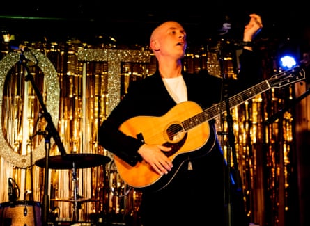 Lekman on stage strumming a guitar and singer on stage next to drums and cymbals, in front of a metallic gold background