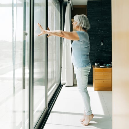 A full-length length of a senior woman with white hair is looking through a window. She is standing on tiptoe in the bedroom.