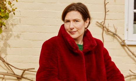 Natasha Solomons wearing a thick dark red coat, sitting in the garden of her home in Dorset, a yellow painted brick wall behind her