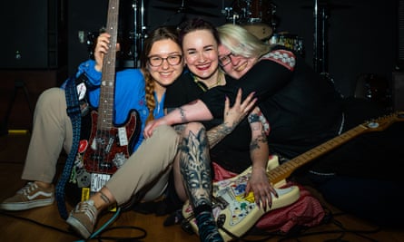 سه زن روی زمین نشسته اند و با لبخندهای بزرگ، دستانشان را دور هم و دو گیتارشان به دوربین نگاه می کنند.