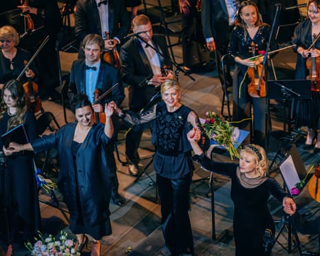 Performers link hands at the front of the stage in front of members of the orchestra