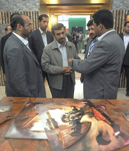 A group of men including Mahmoud Ahmadinejad stand in what looks like a conference room, in front of a table where pictures from the game are displayed.
