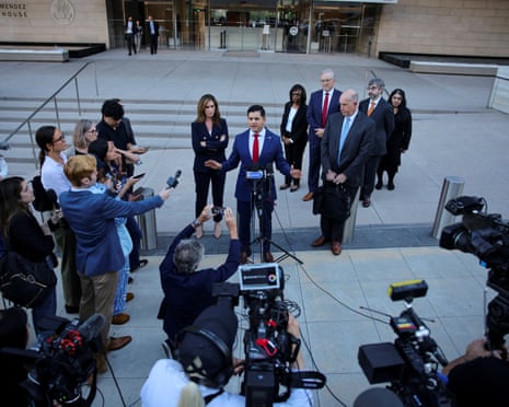 man wearing blue suit and red tie speaks at microphones outside court, surrounded by colleagues and journalists with cameras