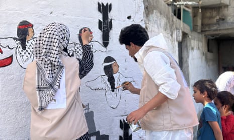 Two artists, one woman, one man, paint images of children and a bomb falling from the sky on a white wall, as two young children watch them work.