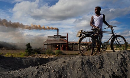 A man with a bicycle stands on a pile of black bullets with a fire roaring in the background