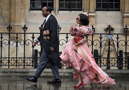 he wears a long black dress coat with medals pinned to his chest and she has an elaborate, floating pink dress with furled petal-style pattern around the neck and a hat; she clutches a bright red handbag and wears glittery red, high-heeled shoes.