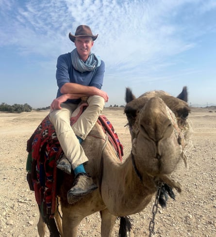 Dan Snow wearing a wide brimmed hat sits on a camel in a desert