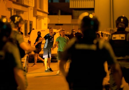 Protesters clash with police in Torre-Pacheco: a man shouts at riot police with his arms outstretched as he stands with others in a street.