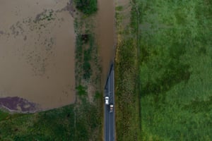 An aerial view of flooding in Geehi, NSW, shows two cars on a road that has become flooded and is surrounded by green fields, some of which are also covered in brown flood waters