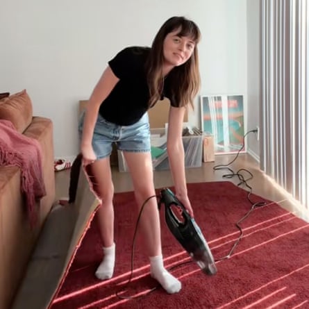 A young female pinch agelong brownish hairsbreadth and a fringe wearing a achromatic t-shirt and denim shorts and achromatic socks stands successful an flat connected a ample portion of reddish carpet holding an handheld vacuum cleaner.