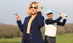 Olga Koch, with a suit jacket but no shirt underneath and holding a cigarette, smiles slightly as she touches her sunglasses as a man in a baseball cap swings a golf club behind her in a field