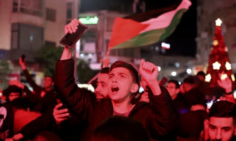 A young man at the front of a crowd raises both hands in the air as someone waves a large Palestinian flag behind him