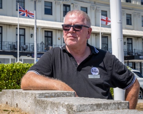 Tom Armstrong stands outside with his arm resting on a wall. A building behind him has the union flag and St George's flag flying