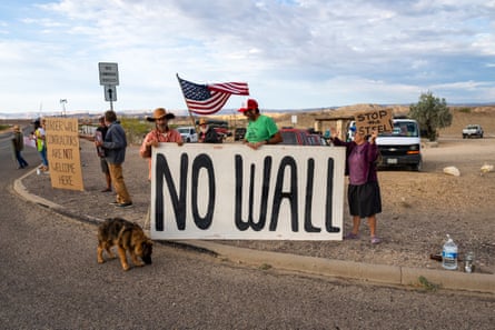 People in a small parking lot amid a flat, desert landscape hold a sign that says ‘No wall.’