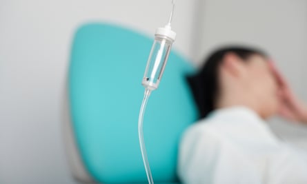 IV drip in foreground, woman in hospital gown with hand on forehead blurry in background.