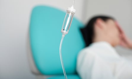 A stock image of an IV tube in the foreground, with a woman resting her hand against her forehead, appearing stressed or in pain, blurred in the background, wearing a hospital gown and reclining on a teal pillow.