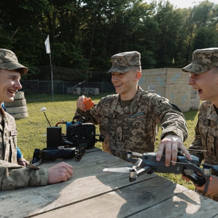 Three young male cadets in camouflage jackets and caps sit around a wooden table with a drone and its control units. They are all laughing and smiling.