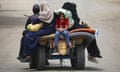 A child sitting on the back of a cart looks downcast as others holding belongings sit on top of mattresses