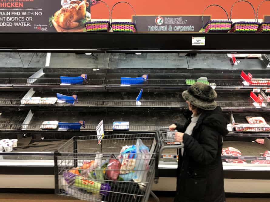 A woman pushes a shopping cart past rows of empty shelves in the meat department of a grocery store.