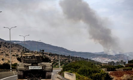 Image of hills with smoke rising from an indistinct point in the distance, with an Israeli tank parked on the road in the foreground
