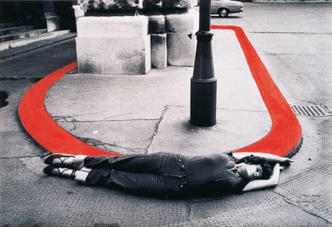 A black and white image of a raised oval section of pavement surrounded by road; in the foreground, a woman lies on her side with arms and legs extended, her body curved against the shape of the raised section; the curb has been colored red in post-production.