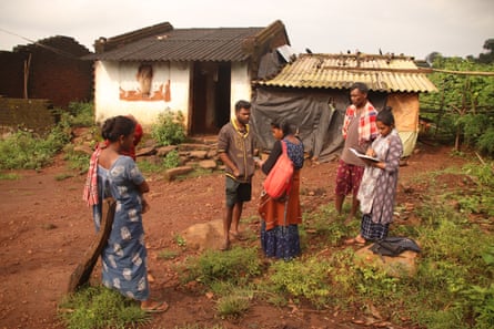 A group of Indian people stand in a farm field outside a shack.