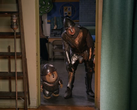 A teddy bear and a young man stand in a doorway wearing knight’s armour