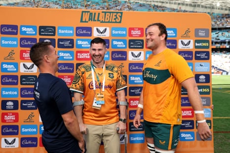 Nathan Charles flips a coin as rugby players Codie Taylor and Harry Wilson watch, smiling