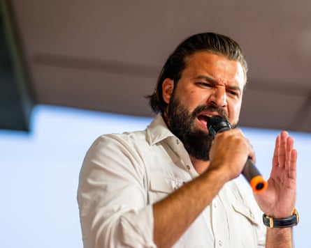 A bearded man speaks into a mic while gesturing with his hand