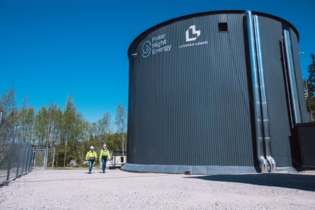 A tall dark grey iron-clad cylinder with silver pipes on one side. It has trees and a blue sky behind it and two people are walking by it wearing white helmets and hi-vis jackets.