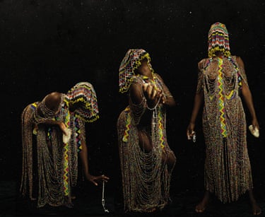 Three figures draped in elaborate beaded garments and headpieces stand against a starry black background