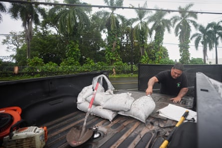 Man pulls at white sandbags in bed of pickup, with line of palm trees behind him under cloudy skies.