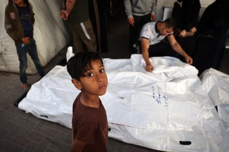 A young boy looks towards the camera as a man crouching behind him appears to open one of various bulging body bags.