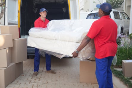 Two removal men in red T-shirts and blue trousers and baseball caps lift a cream sofa out of a van.