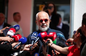 Edi Rama, wearing round red-rimmed sunglasses, speaks into microphones held up by journalists