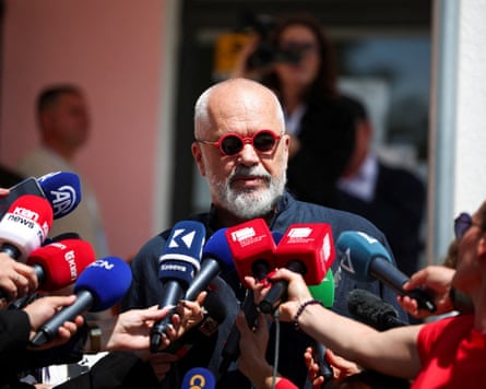 Edi Rama wearing red sunglasses surrounded by reporters holding microphones