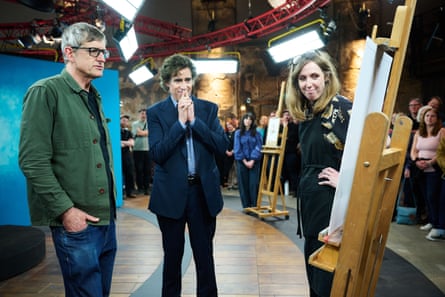 A man in a green jacket with grey hair and glasses and a man with curly brown hair wearing a dark suit clasping his hands in front of his face look at a picture on an easel with a woman smiling standing next to it