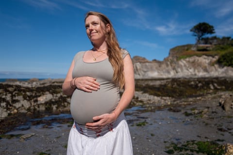A pregnant woman, with her hands on her ‘bump’, stands on a beach and looks off into the distance. qhxidiqxkiqrrinv