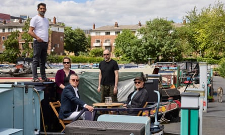 Four men and a woman gathered on the deck of a dark blue steel narrowboat, sitting around a table or standing behind; one man is standing on the cabin roof. Other moored boats and electricity hook-up points can be seen with trees and buildings in the background across the other side of the canal 