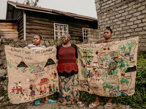 An older African woman stands outside a house as two younger women hold two large embroidered artworks on either side of her