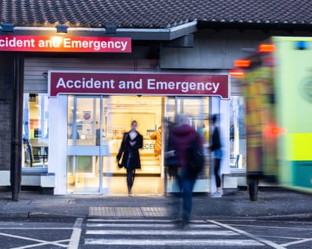 Blurred people walk past a lit Accident and Emergency hospital entrance at night