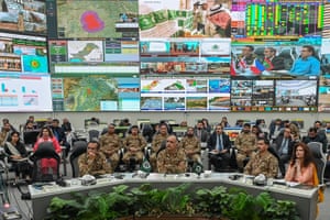Officials and others in military uniform sit in a room with a large display of monitors above them