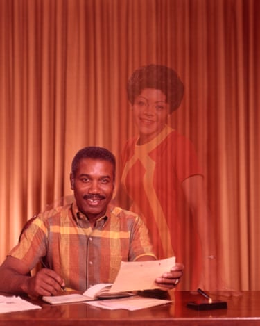 Colour photograph of a man sat on a desk with a pen and paper in hand. The photo has a hologram of a woman in view.