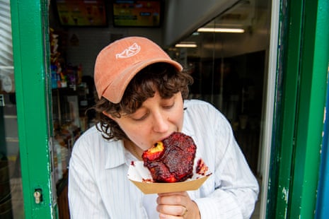A woman wearing a pink hat and a light blue shirt biting into a pineapple covered in red sauce